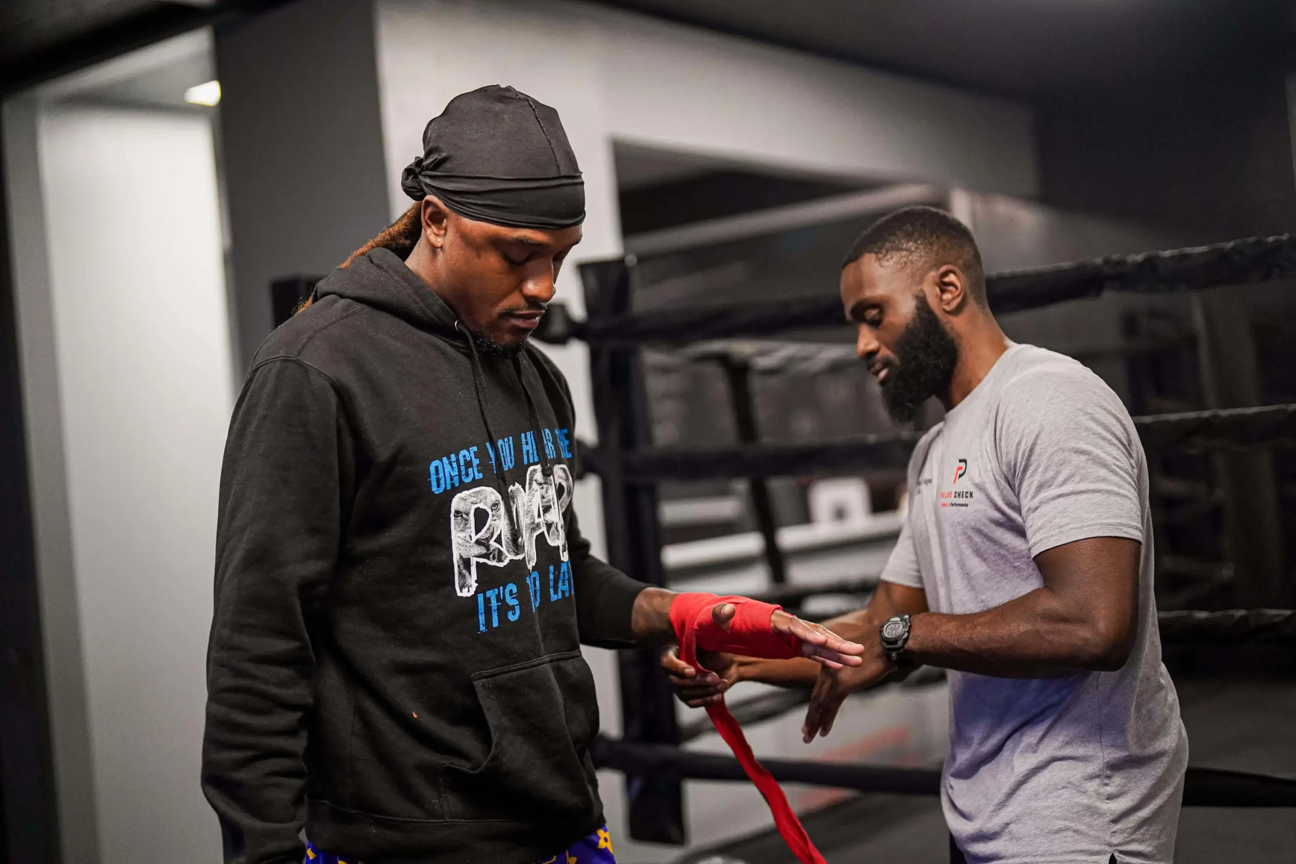 Pulse Check coach wrapping a boxer's hand with red hand wraps beside the boxing ring before training